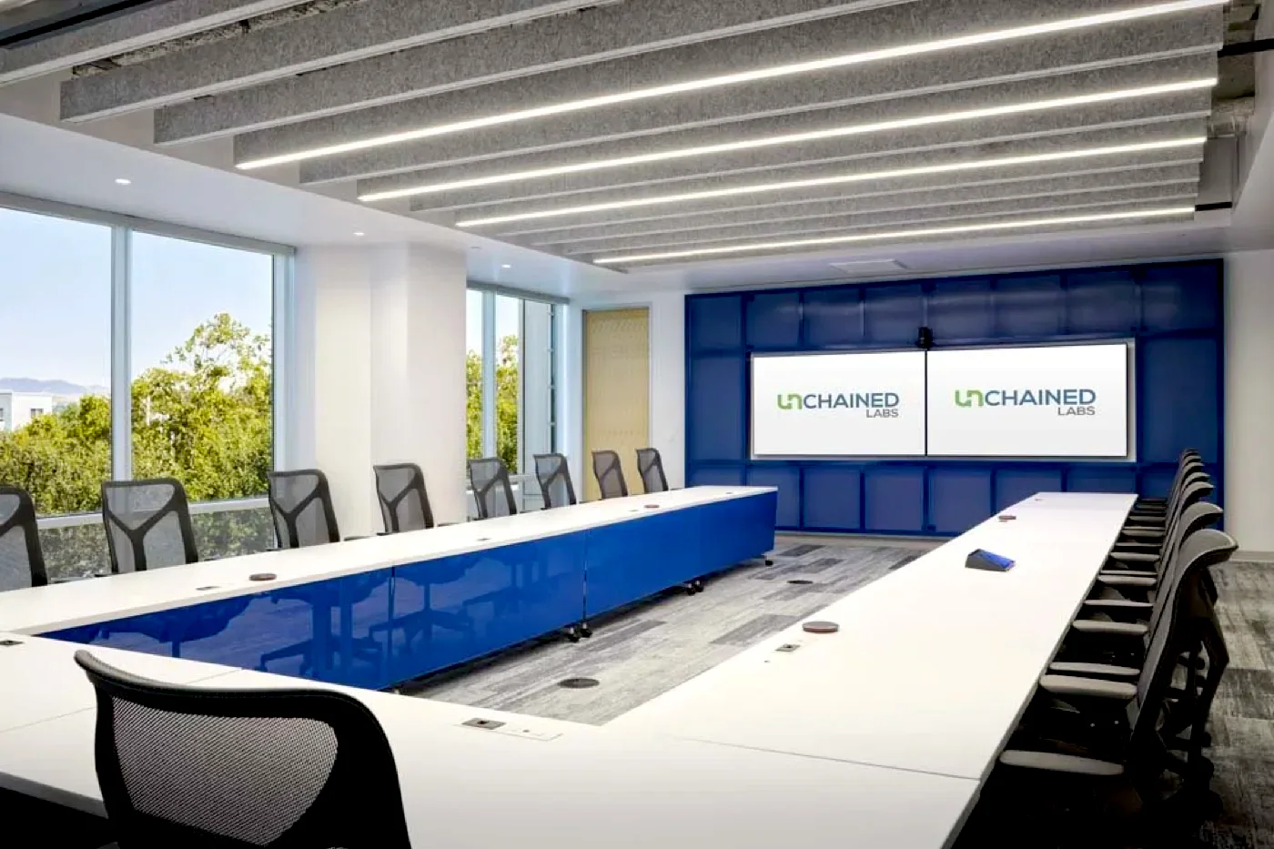 Conference room with a long blue table, linear ceiling lighting, and two wall displays